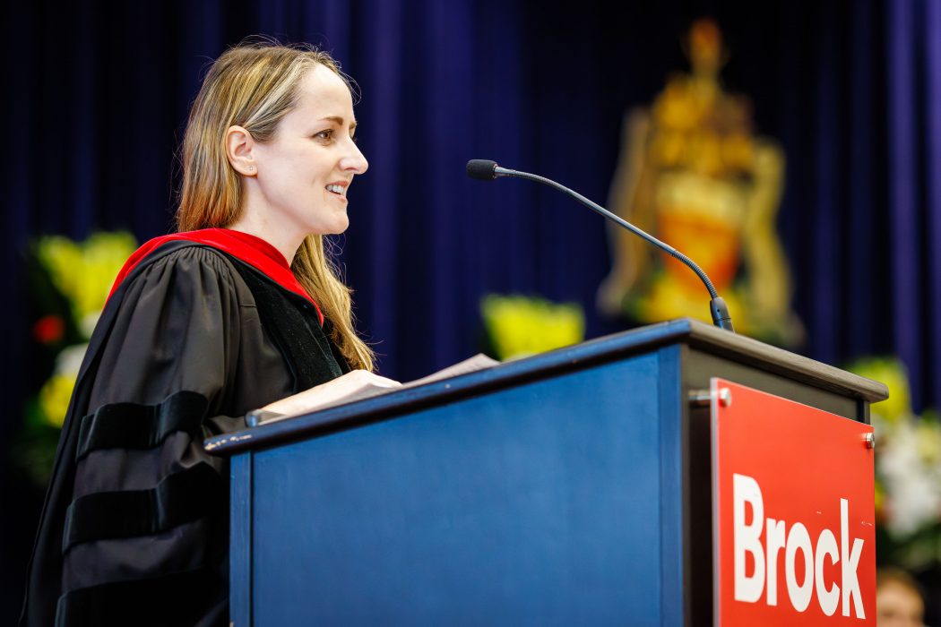 A woman in academic robes speaks at a podium during a graduation ceremony.