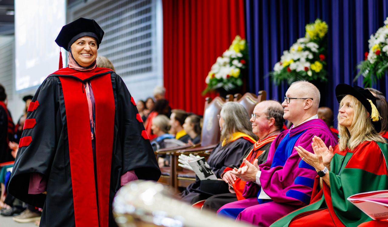 A woman in academic regalia smiles and walks across the stage during a university graduation ceremony.