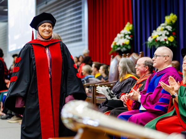 A woman in academic regalia smiles and walks across the stage during a university graduation ceremony.