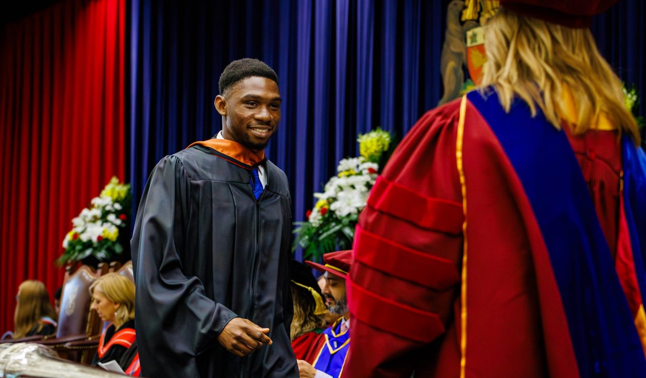 A young man in academic robes walks across the stage during a university graduation ceremony.