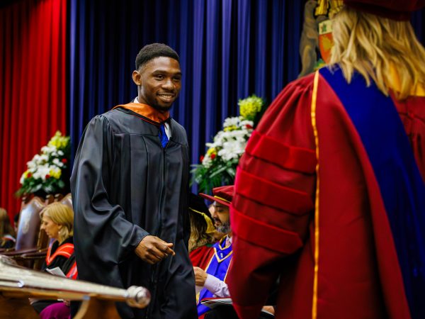 A young man in academic robes walks across the stage during a university graduation ceremony.
