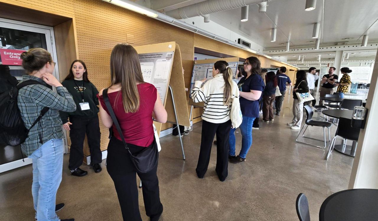 People look at research posters set up on easels in a hallway while speaking with one another.