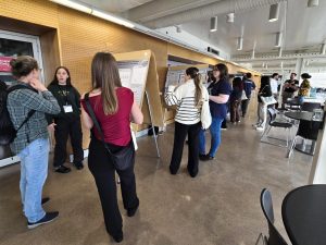 People look at research posters set up on easels in a hallway while speaking with one another.