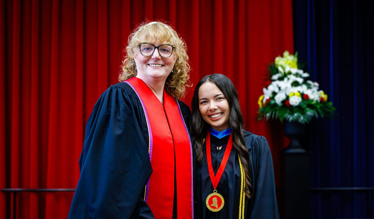 Two women in academic robes pose for a photo on stage at a university graduation ceremony.