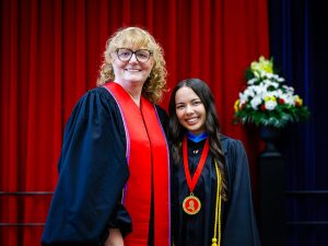 Two women in academic robes pose for a photo on stage at a university graduation ceremony.