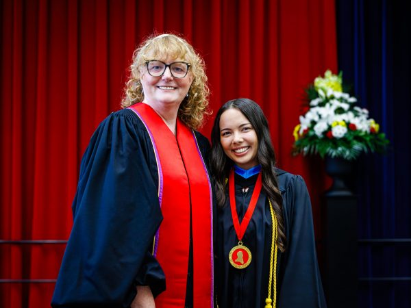 Two women in academic robes pose for a photo on stage at a university graduation ceremony.