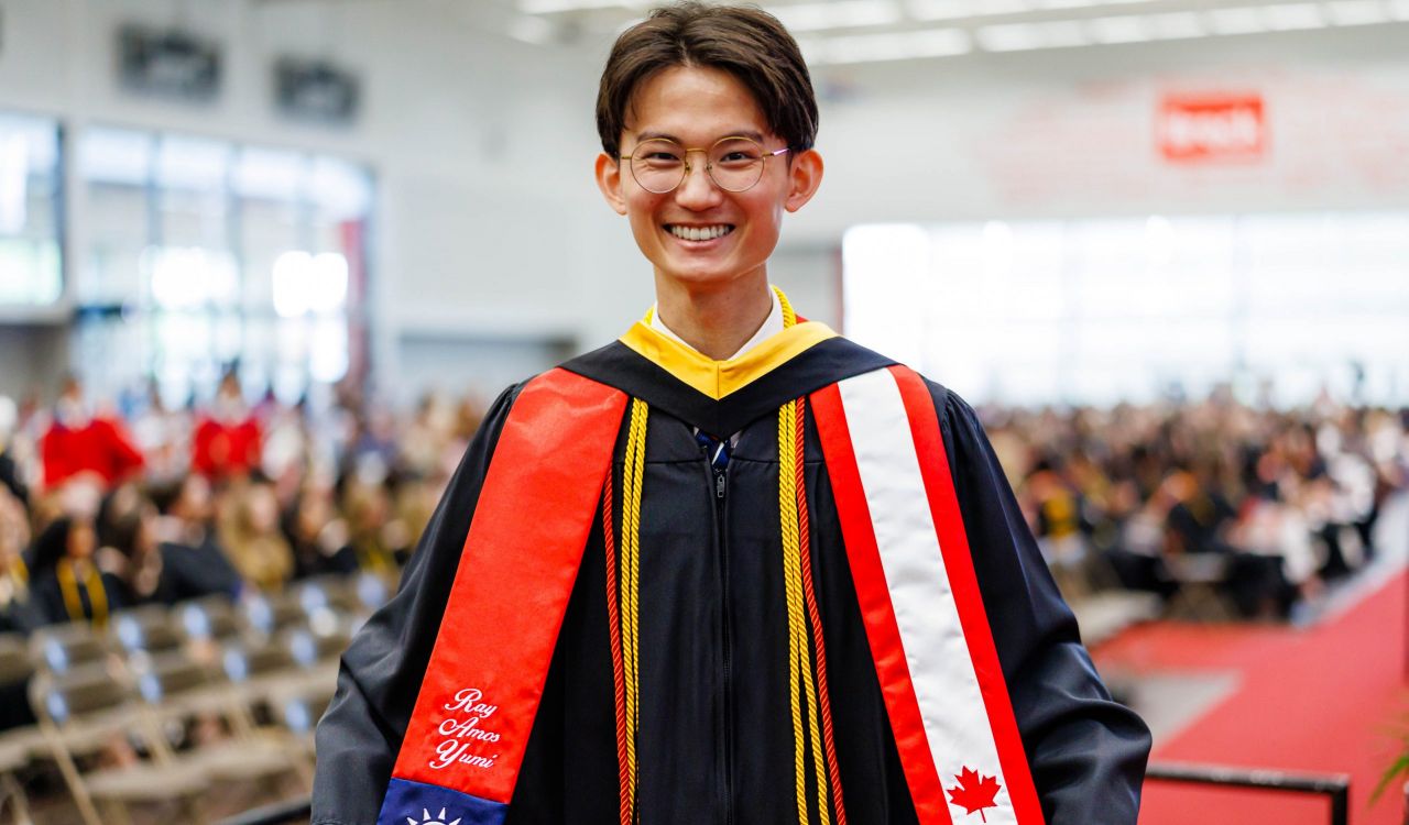 A graduate wearing ceremonial robes smiles in front of an audience at a Convocation ceremony.