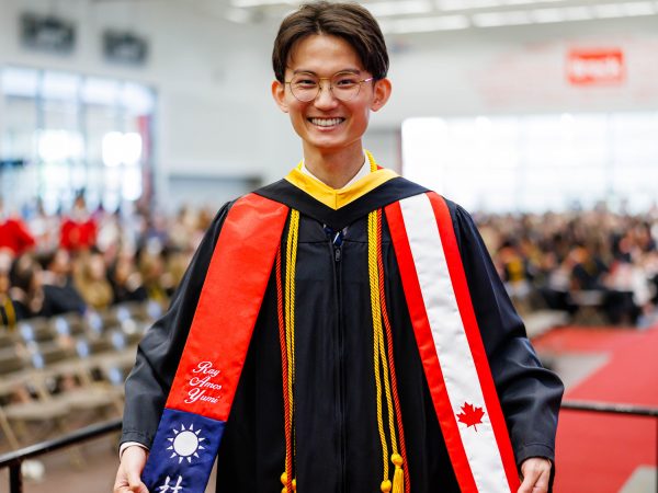 A graduate wearing ceremonial robes smiles in front of an audience at a Convocation ceremony.