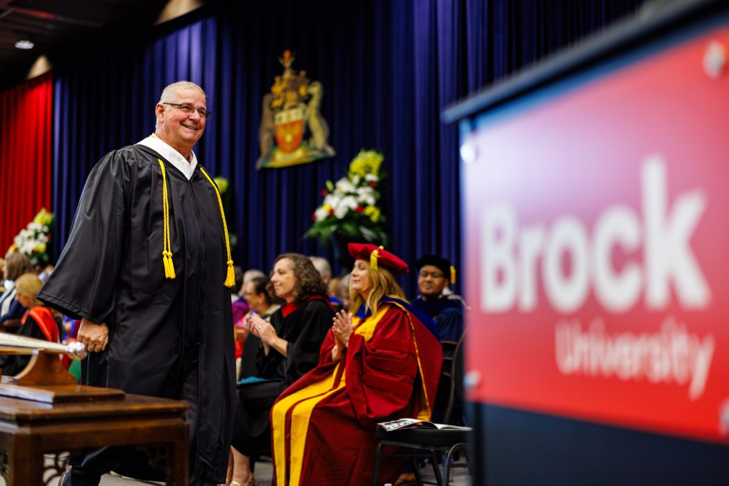 A man in a Convocation gown walks across a stage filled with people in regalia sitting on chairs.