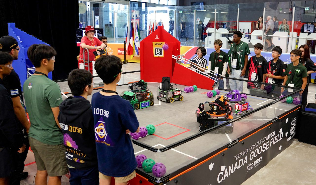 Teams young students gather around a competition ring controlling robots with remote controls.
