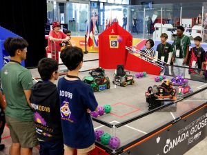 Teams young students gather around a competition ring controlling robots with remote controls.