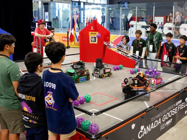 Teams young students gather around a competition ring controlling robots with remote controls.