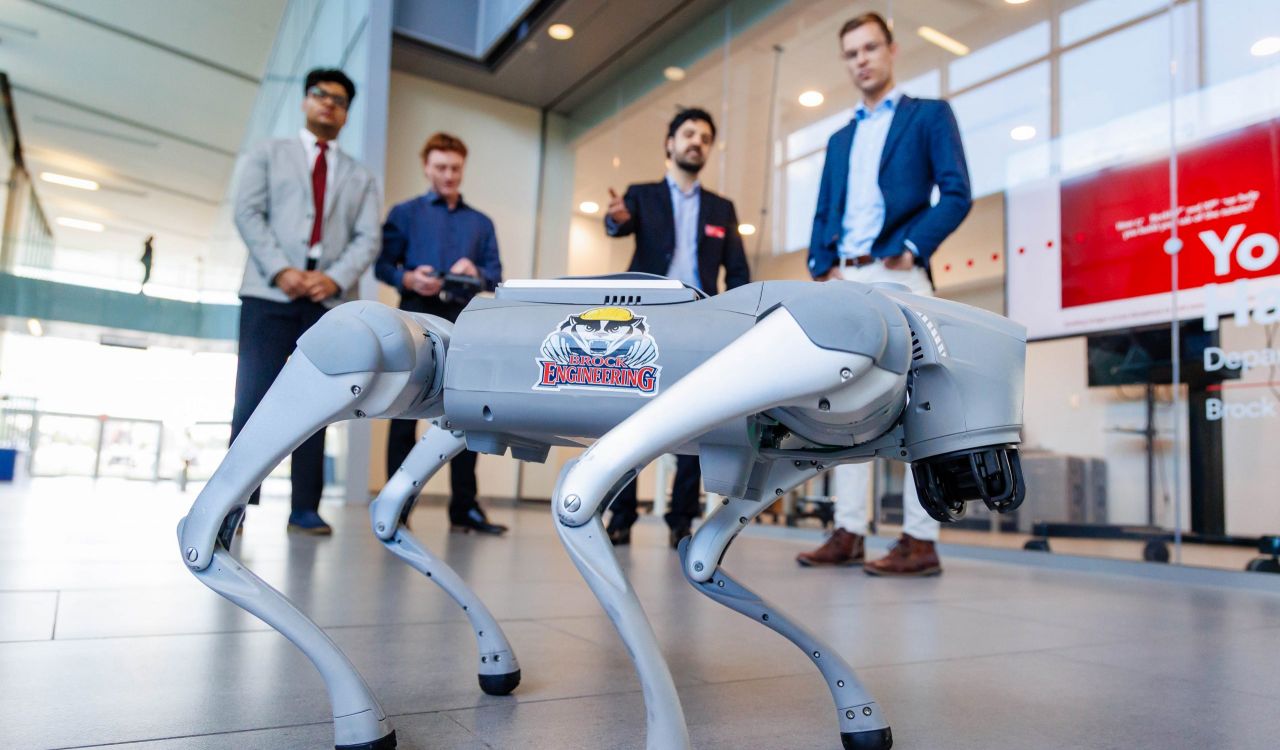 Four people stand behind a robot dog featured prominently in the foreground. The dog has a Brock Engineering sticker on it with an image of a badger wearing a hard hat.