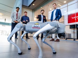 Four people stand behind a robot dog featured prominently in the foreground. The dog has a Brock Engineering sticker on it with an image of a badger wearing a hard hat.