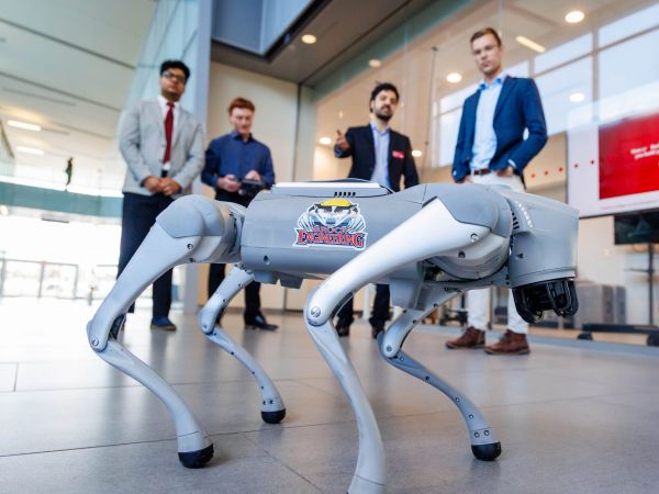 Four people stand behind a robot dog featured prominently in the foreground. The dog has a Brock Engineering sticker on it with an image of a badger wearing a hard hat.