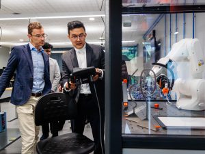 Two people look over a robotic arm in a case. One person is holding a remote control.