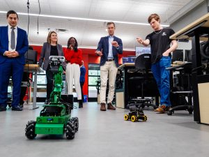 Four people stand together looking over a robotic with wheels on the floor.