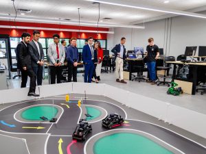 A group of people stand together looking over a miniature robot race track with self-driving cars.