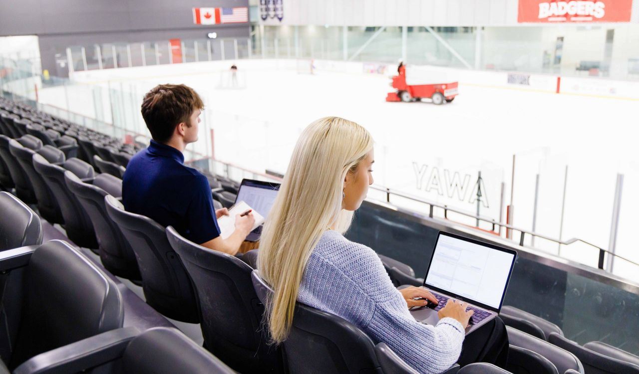 Two students work on laptops in the stands of an ice rink.