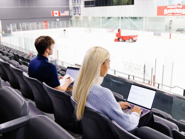 Two students work on laptops in the stands of an ice rink.