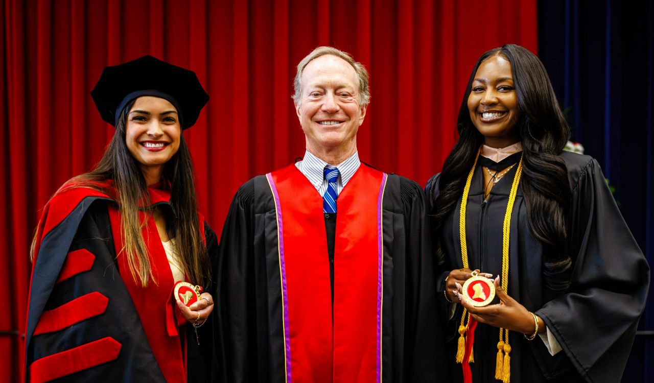 Three people in academic regalia smile for a photo in front of a red curtain. The two people on either side hold red and gold medals.