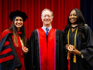 Three people in academic regalia smile for a photo in front of a red curtain. The two people on either side hold red and gold medals.