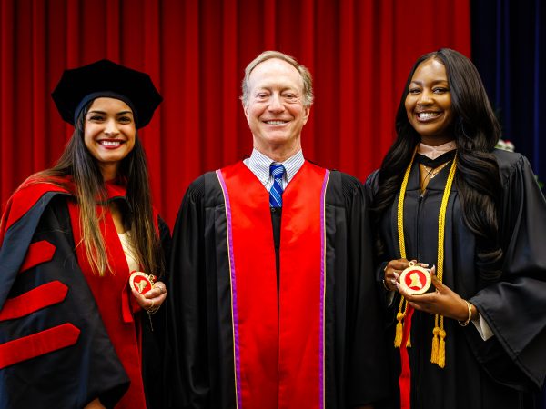Three people in academic regalia smile for a photo in front of a red curtain. The two people on either side hold red and gold medals.