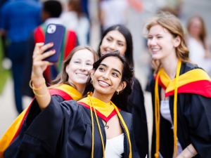 Four young women in university graduation gowns pose for a selfie.