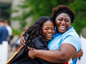 Two women, one in a convocation gown, embrace outside.