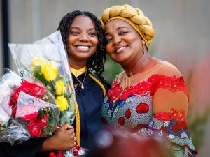 Two women stand together, one in a Convocation gown holding flowers.