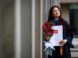 A woman in a Convocation gown stands outside holding roses and a Brock-branded folder.