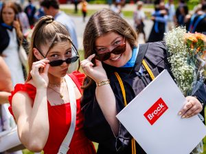 Two women tip their sunglasses while smiling. One is wearing a Convocation gown and holding a Brock University-branded folder.
