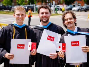 Three men in Convocation gowns stand together outside holding Brock University-branded folders.