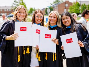 Four women in in Convocation gowns stand together outside while holding Brock University-branded folders.