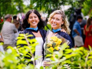 Two women graduates in Convocation gowns stand together outside.