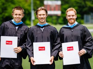Three men graduates in Convocation gowns stand together outside while holding Brock University-branded folders.