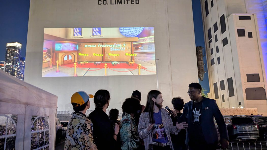 A group of people talk to a person holding a microphone in front of a building with a video game projected onto it.