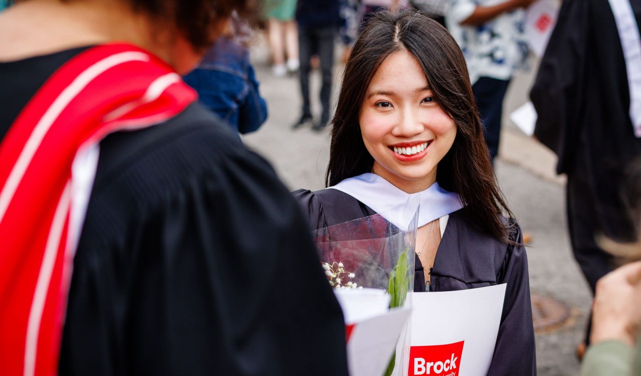 A graduate in a Convocation gown smiles while holding flowers and a Brock University branded folder.