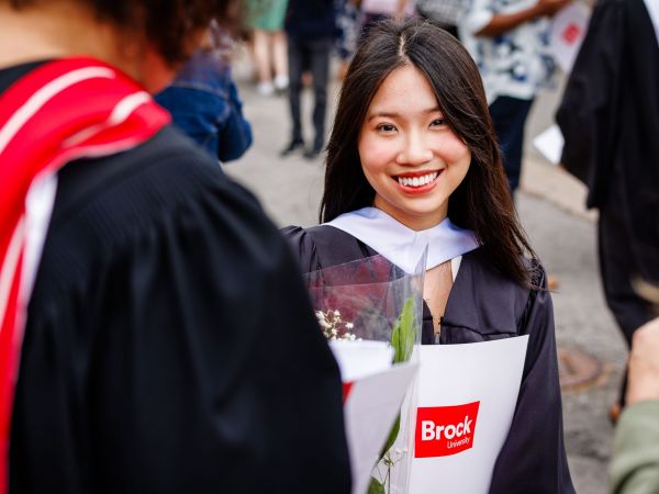 A graduate in a Convocation gown smiles while holding flowers and a Brock University branded folder.