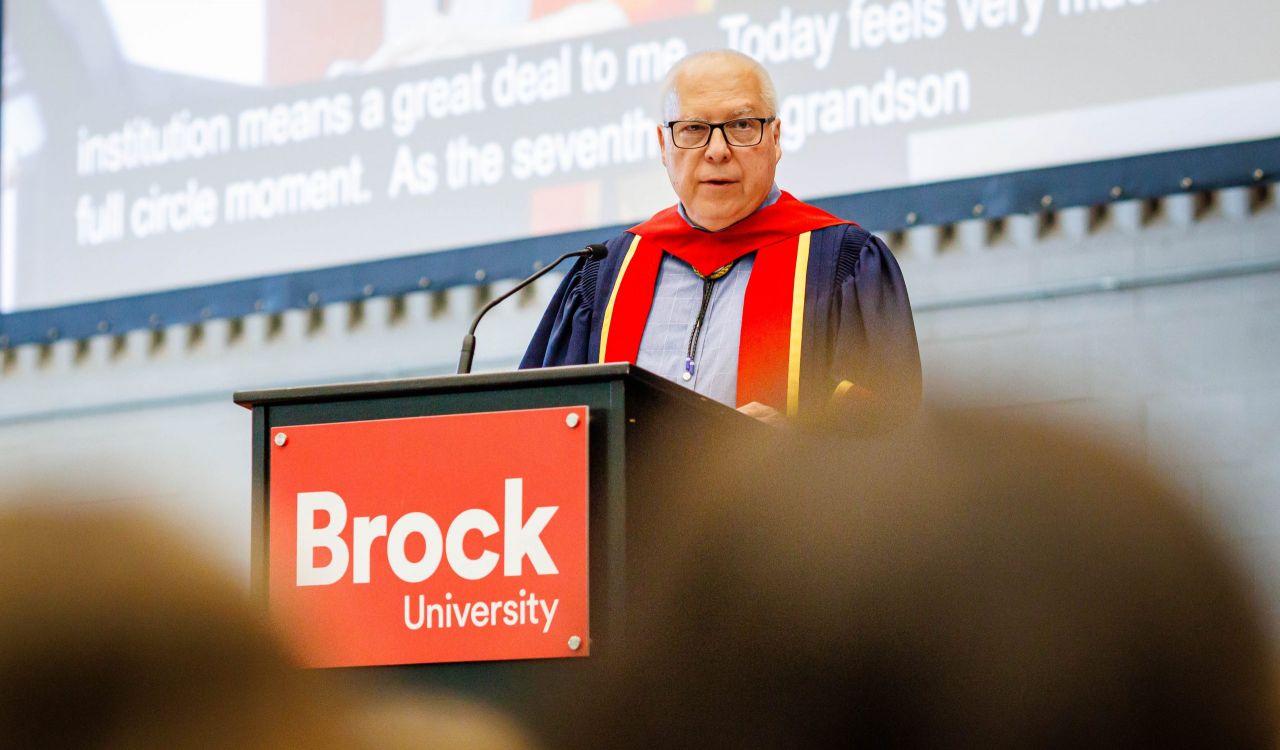 A man in a Convocation gown stands at a Brock University branded podium.