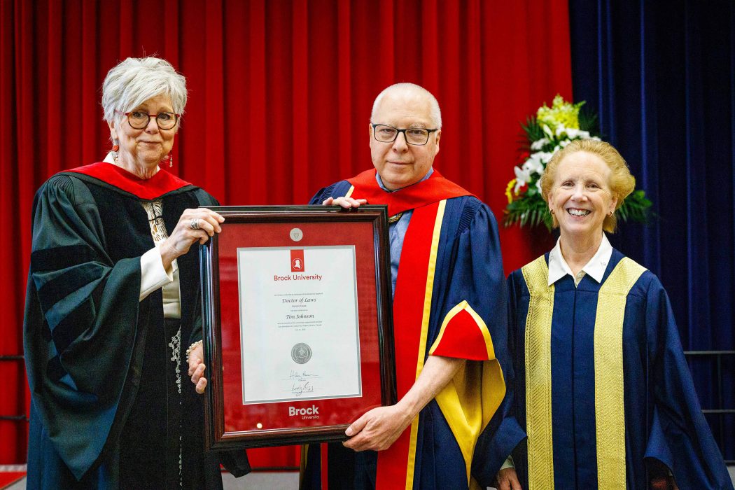 A man receives a framed degree from two woman. All are wearing Convocation regalia.