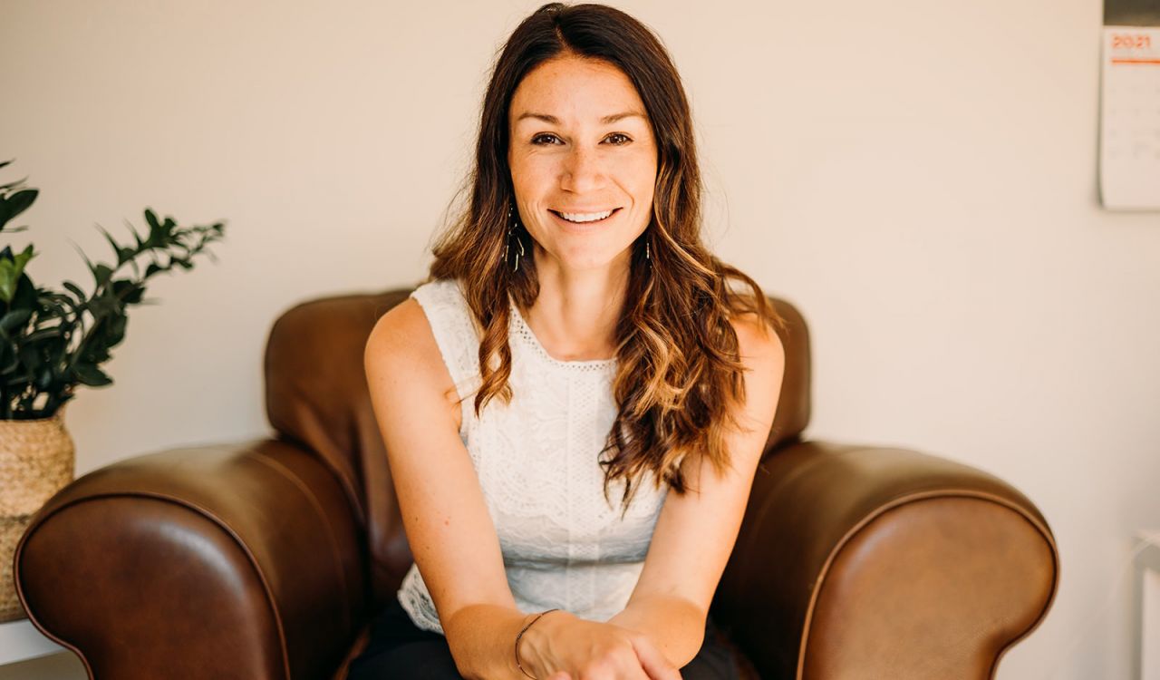 A smiling woman sits on a chair in front of a beige wall.