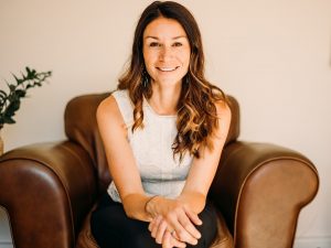 A smiling woman sits on a chair in front of a beige wall.