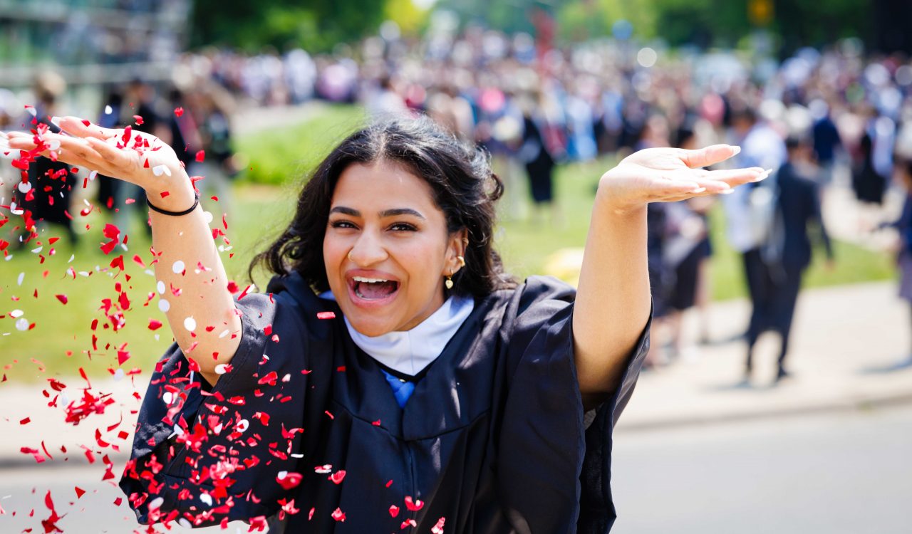 A young woman in academic robes throws confetti at a university graduation ceremony.
