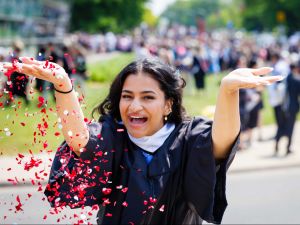 A young woman in academic robes throws confetti at a university graduation ceremony.