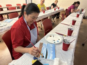 In a classroom setting, a woman sits at a long communal table and smiles as she paints a small waterfront scene. There are four other women in the room, also sitting at long tables and paining.