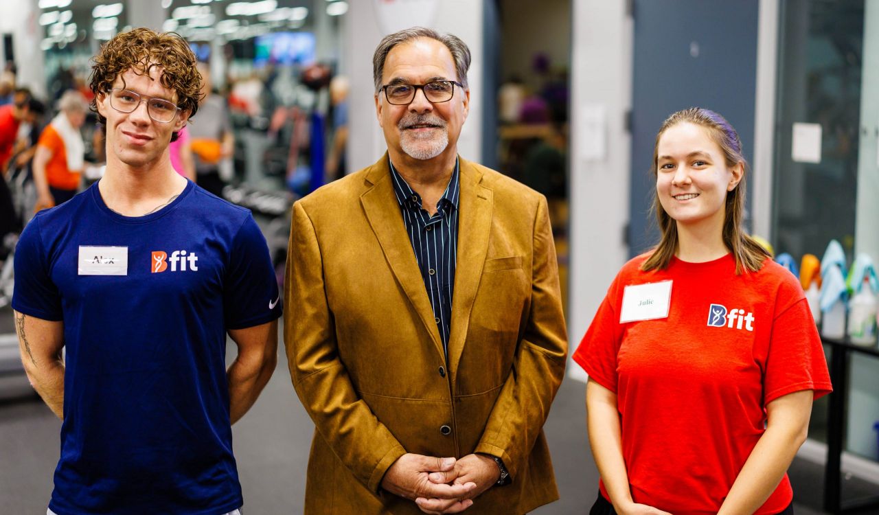 A man standing with 2 students in a gym where people are exercising.