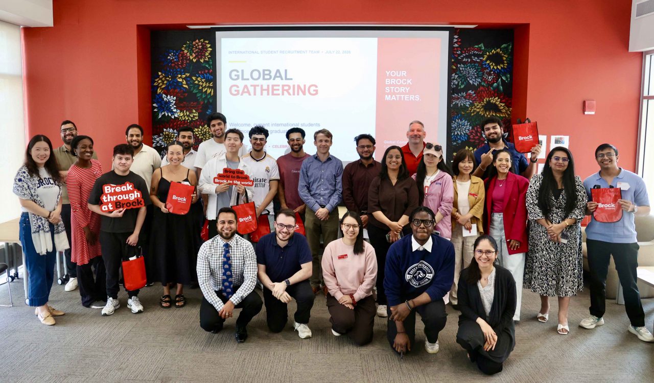 A large group of people pose for a photo in front of a red wall at Brock University.