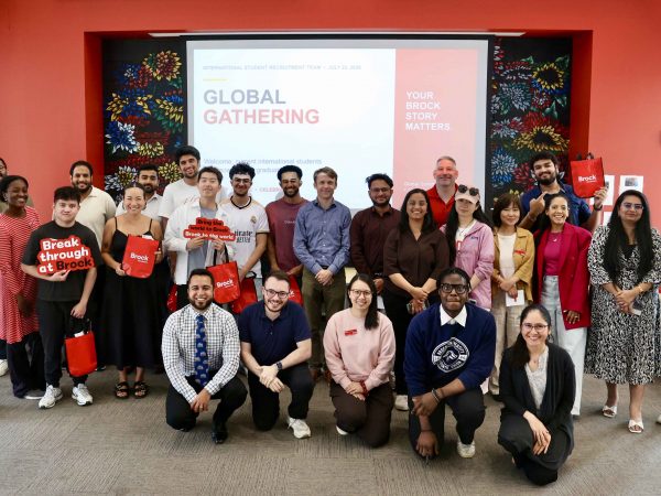 A large group of people pose for a photo in front of a red wall at Brock University.