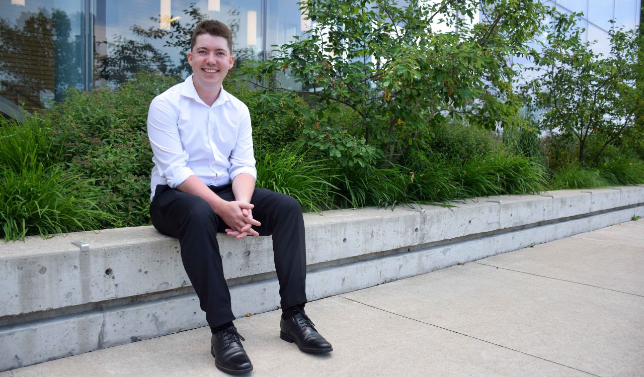 A young man sits on a ledge in front of a building with a raised garden behind him.